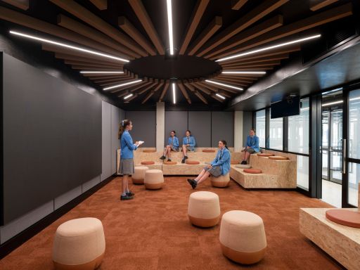 Group of people in blue uniforms gathered in a modern lounge with round beige ottomans and low tables under a radial wooden ceiling light fixture.