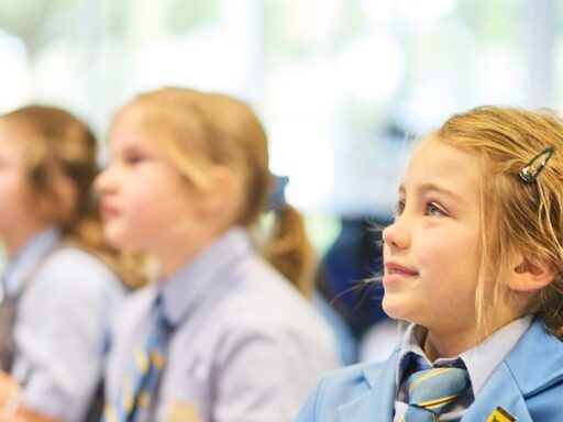 Girl in a blue blazer with a striped tie, sitting in a classroom with other students blurred in the background, looking to the right.