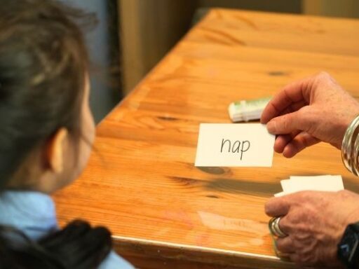 Child sits at a wooden table while an adult places a flashcard reading 'nap' on the surface, suggesting a learning or reading activity.
