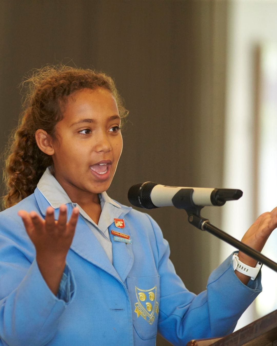 Young girl in a blue school blazer speaks into a microphone at a podium during a school event.