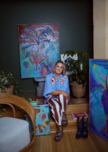 Woman sits on a wooden step among vibrant abstract paintings and potted plants in an art studio.