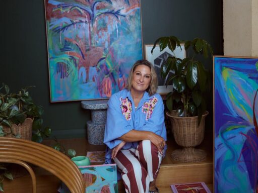 Woman sits on a wooden step among vibrant abstract paintings and potted plants in an art studio.