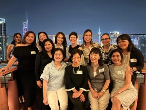 Thirteen women in business-casual attire pose together on a nighttime rooftop terrace, with city lights and skyscrapers in the background.