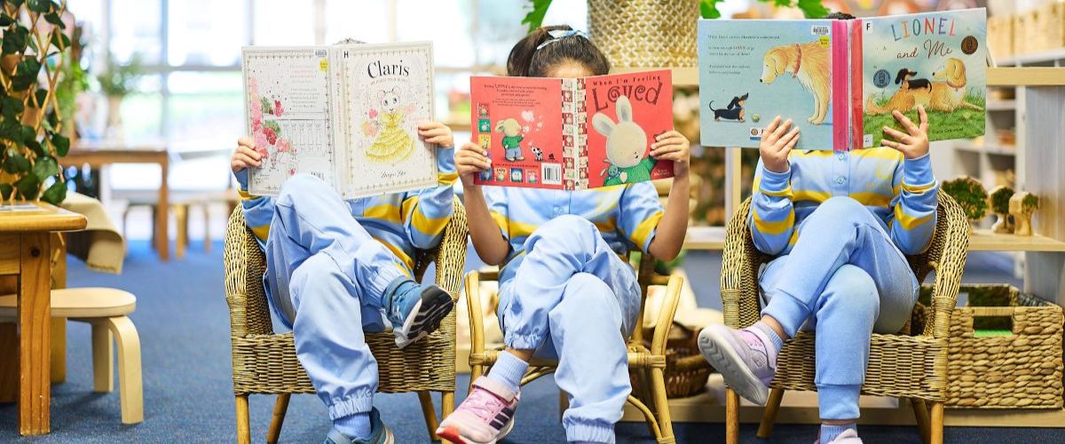 Three children in matching blue outfits sit in wicker chairs reading colorful picture books in a bright library setting.