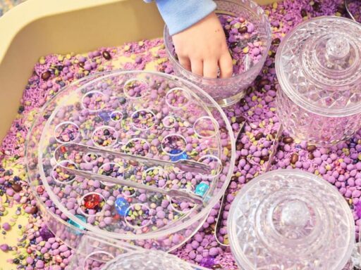 Child's hands scoop pink/purple beads from a crystal-topped sensory tray with clear jars nearby.