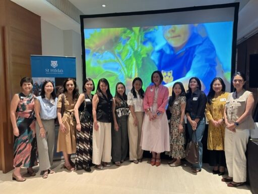 Group of women posing for a photo in a conference room, with a large colorful screen behind and a St Hilda's banner on the left.