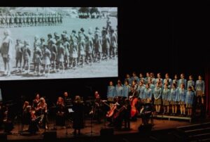 Choir in blue dresses with an orchestra on stage; a black-and-white photo of marching children is projected behind them.
