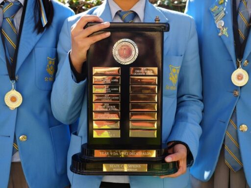 Student in a blue blazer holds a tall award plaque with gold nameplates from a school competition.