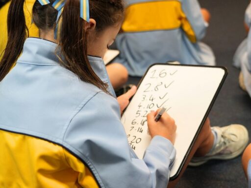 Teen student in a blue and yellow uniform writing on a whiteboard tablet while seated on the floor in a classroom group activity.