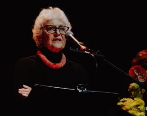 Older woman with white hair and glasses speaking at a podium on a dimly lit stage, wearing a black outfit and red necklace.