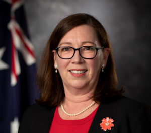 Professional headshot of a woman with glasses, pearl necklace, and red blouse, wearing a red flower pin; dark blazer and flag backdrop behind her.