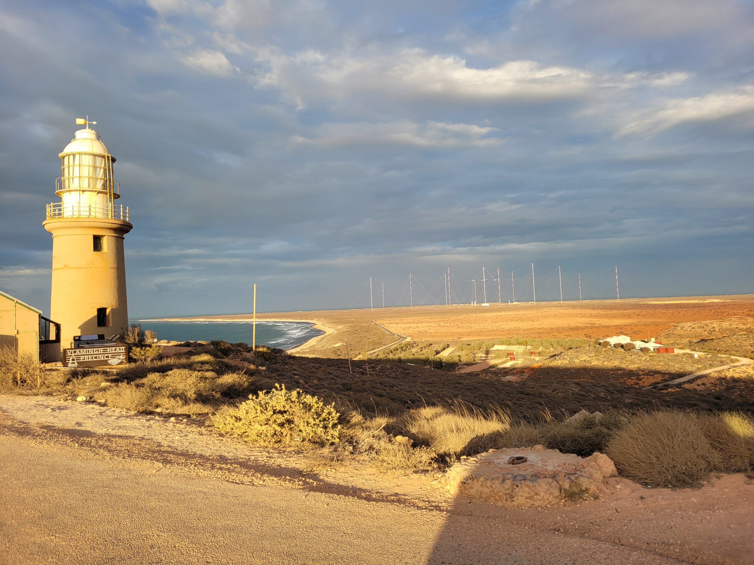 Harold E Holt Naval Communications Station - St Hilda's Anglican School For Girls White lighthouse on a sandy coastal landscape with a calm sea and cloudy sky to the right.