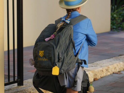 Child with a large gray backpack walking on a sidewalk, wearing a blue jacket and straw hat with a blue ribbon.