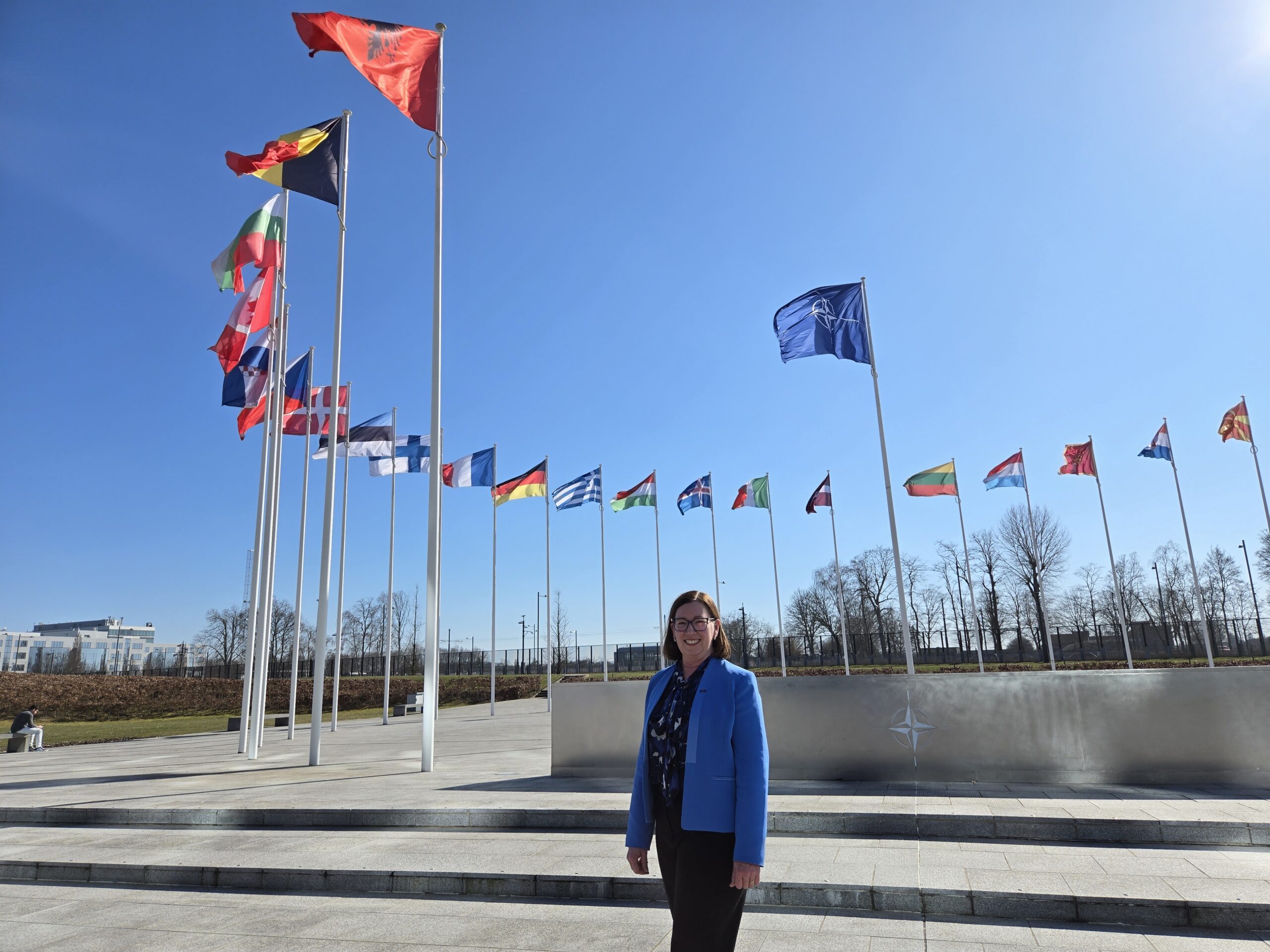NATO HQ - St Hilda's Anglican School For Girls Smiling woman in a blue blazer stands in front of a row of tall flagpoles with international flags against a clear blue sky.