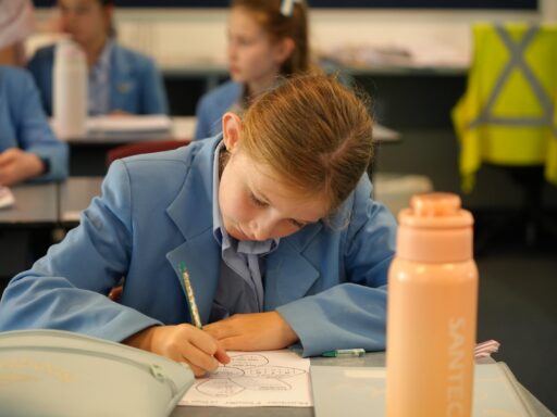 Girl in a blue school uniform writes on a worksheet at a desk, pink water bottle in the foreground, classroom in the background.
