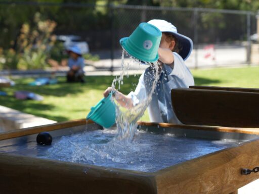 Child with light blue hat tilting a turquoise bucket over a wooden fountain, water spraying into the basin in a sunny park.