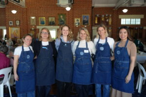 Six women in blue denim aprons stand side by side smiling in a brick-walled room with portraits on the wall behind them, likely volunteers at an event.