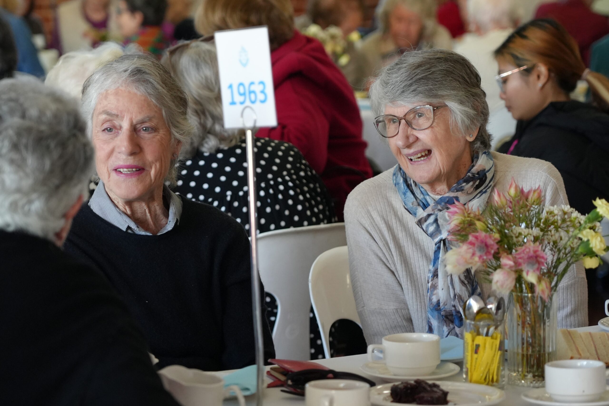 STH08834 - St Hilda's Anglican School For Girls Two elderly women sit at a table with a '1963' table sign, chatting while a bouquet and plates are on the table.