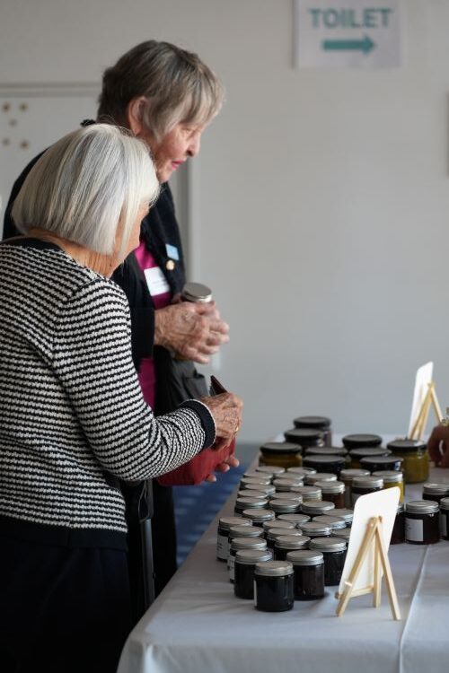 STH08958 - St Hilda's Anglican School For Girls Two elderly women stand at a table of small jars with dark lids, examining preserves at a market or booth.