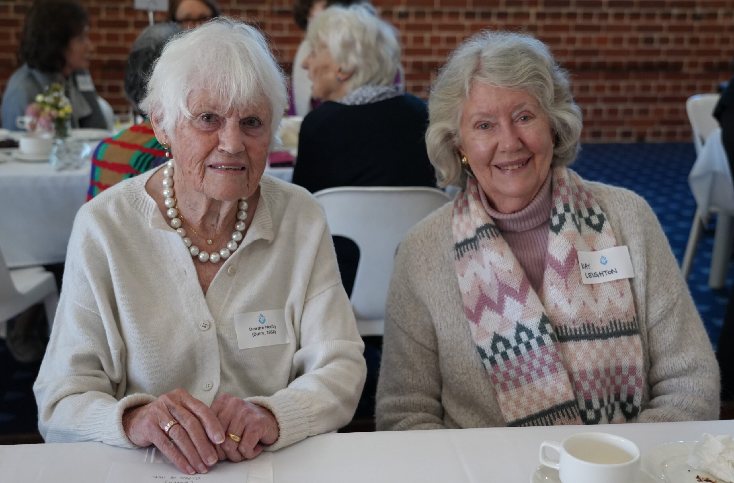 STH09023 - St Hilda's Anglican School For Girls Two elderly women sit together at a table during a social event, smiling at the camera; one wears a pearl necklace and white sweater, the other a patterned scarf.