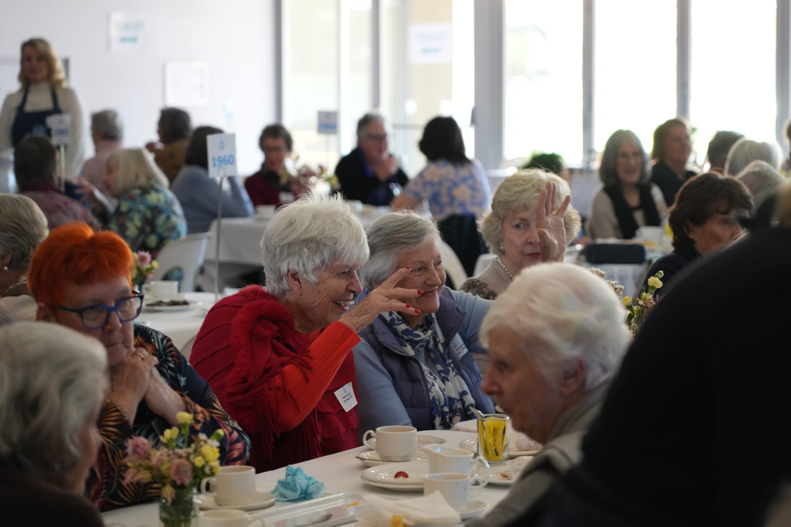 STH09069 - St Hilda's Anglican School For Girls Elderly adults at a luncheon, seated around tables with cups, plates, and small flower arrangements in a bright room.
