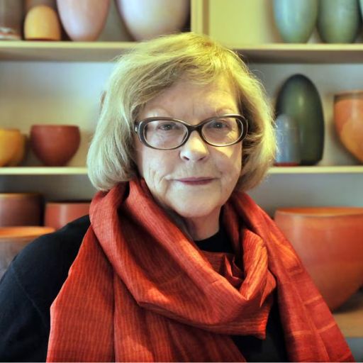 Portrait of a woman with short gray hair and glasses, wearing a red scarf, standing in front of shelves filled with colorful pottery bowls and vases.