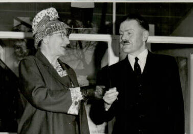 Una Mitchell and Library Opening Mr F A Sharr State Librarian - St Hilda's Anglican School For Girls Black-and-white photo of an elderly woman in a patterned hat and coat shaking hands with a man in a dark suit and tie at a doorway.