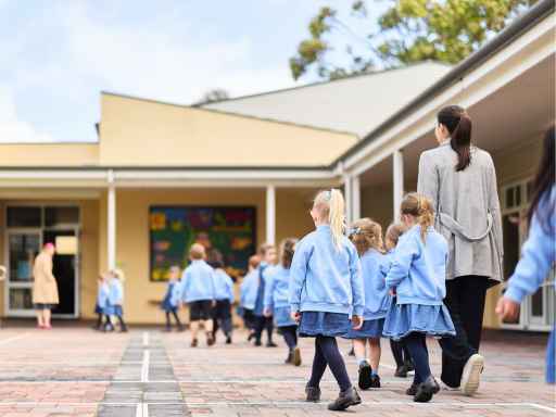 Teacher supervising a line of young students in blue uniforms as they walk outside a school building on a sunny day.
