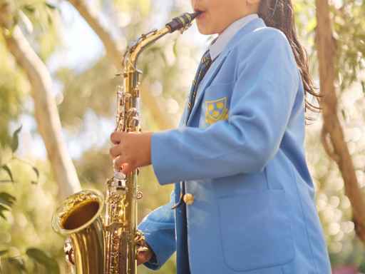 Girl in a blue school blazer playing a saxophone outdoors in a sunny park setting