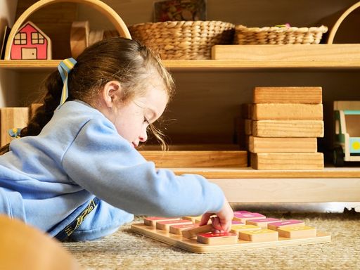 Girl lying on the floor, concentrating on a wooden block puzzle, with shelves of wooden toys in the background.