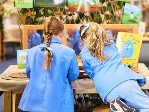 Two girls in blue school uniforms lean over a table to view a display of colorful artwork and cards at a school event