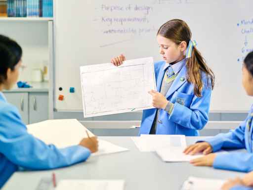 Girl in a blue uniform holds up a chart while classmates look on in a classroom.