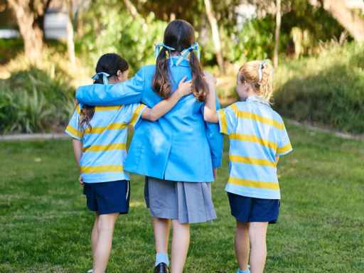 Three children walk arm-in-arm away from the camera in a sunny park, wearing blue and yellow uniforms.