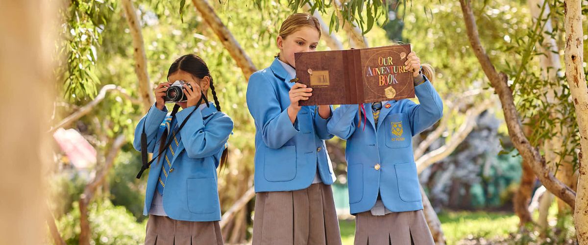 Three girls in blue school uniforms outdoors, one taking a photo while another reads a large adventure book between them.