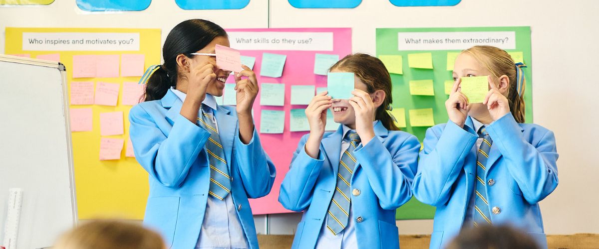 Three students in blue blazers and striped ties hold sticky notes over their eyes in a colorful classroom display.