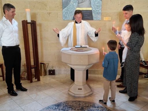 Priest at a baptismal font performing a ceremony; family nearby holds lit candles, with a small child facing the priest.