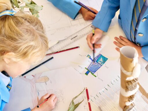 Students in blue uniforms drawing at a table with pencils, rulers, and sketches on large sheets of paper