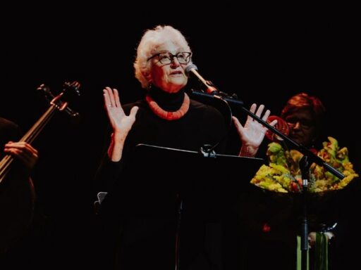 Older woman with white hair and glasses, wearing black with a red necklace, speaking into a microphone on a dark stage, hands raised in emphasis.