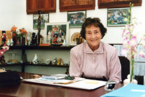 Smiling elderly woman in a pink blouse sits at a desk surrounded by framed photos and shelves with trinkets and ornaments.