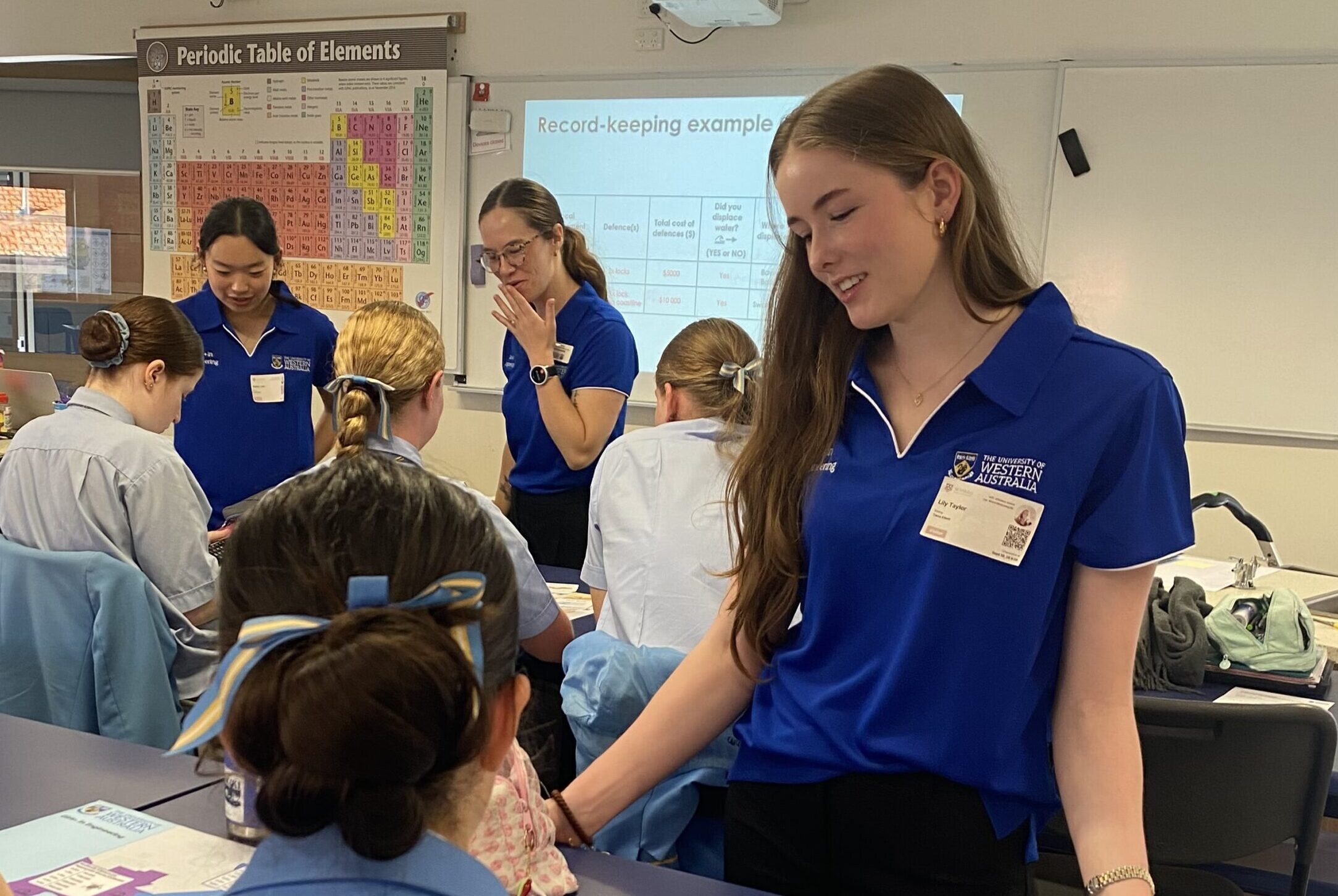 IMG_1316 - St Hilda's Anglican School For Girls Group of students in blue uniforms gathered around desks in a classroom, with a periodic table poster on the wall and a projector screen in the background.