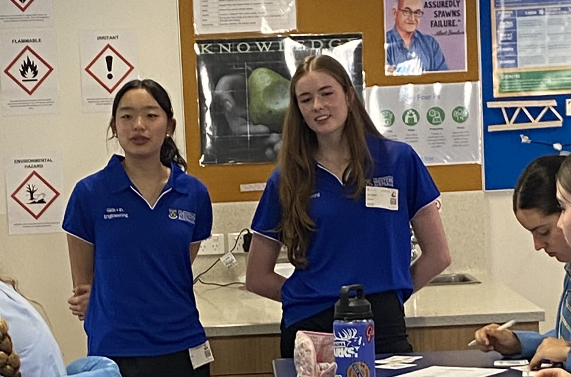 IMG_1318 - St Hilda's Anglican School For Girls Two female students wearing blue polo shirts stand at a desk in a classroom, with posters and papers on the wall behind them.