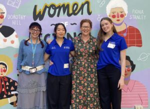 Four women stand together in front of a colorful mural featuring illustrated female portraits and the word 'Women'.