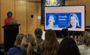 Woman in red sweater speaks at a podium in a wood-paneled room; a large screen displays 'Build New Habits' and 'Atomic Habits' slide.