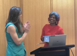 Two women at a podium during a presentation; one in teal with hands together, the other in red smiling at a laptop on the podium against wooden panels.