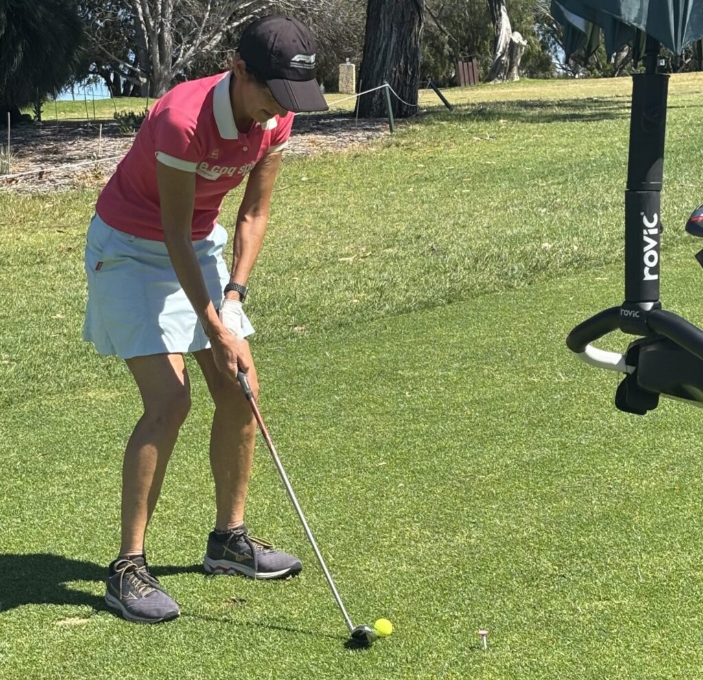 IMG_8664 - St Hilda's Anglican School For Girls Female golfer in a pink polo shirt and white skirt lining up a putt on a sunny golf course with a yellow ball nearby.