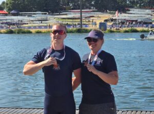 Two rowers posing with medals by a lake; boats stacked in the background, a rowing venue behind them to the shore.