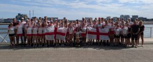 Large group of athletes in white and red uniforms posing by a harbor with England flags displayed.