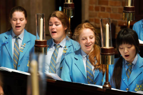 Slide 2 Emily Kempin at School singing - St Hilda's Anglican School For Girls Group of students in blue blazers singing from sheet music in a church choir, with candles nearby.