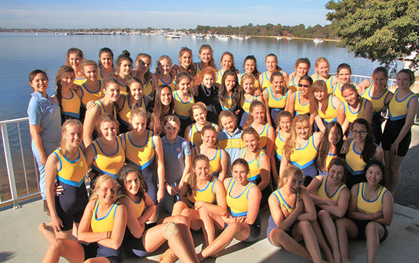 Slide 3 Emily Kemptin St HIldas Rowing 2 - St Hilda's Anglican School For Girls Large group of young women in yellow and blue uniforms posing for a team photo by a lake with boats in the background.