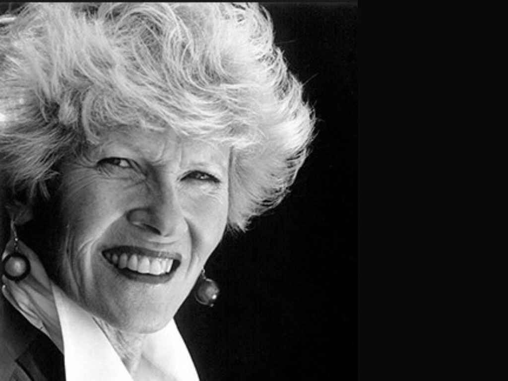 Close-up portrait of a smiling elderly woman with short white hair, wearing pearl earrings against a dark background.